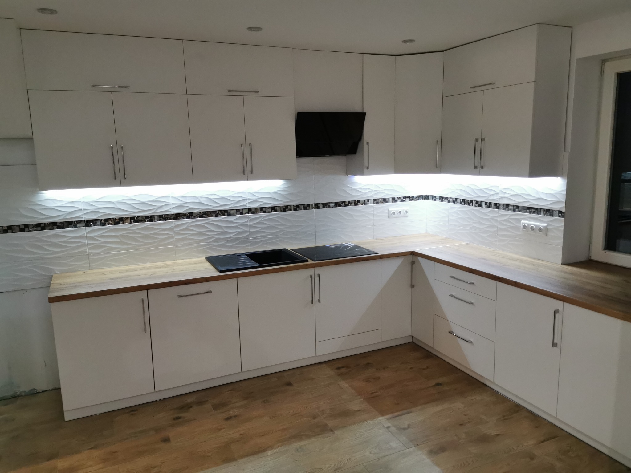 Nowoczesna, biała zabudowa kuchenna z drewnianym blatem, oświetleniem LED podszafkowym i dekoracyjnymi płytkami ściennymi z motywem fal.