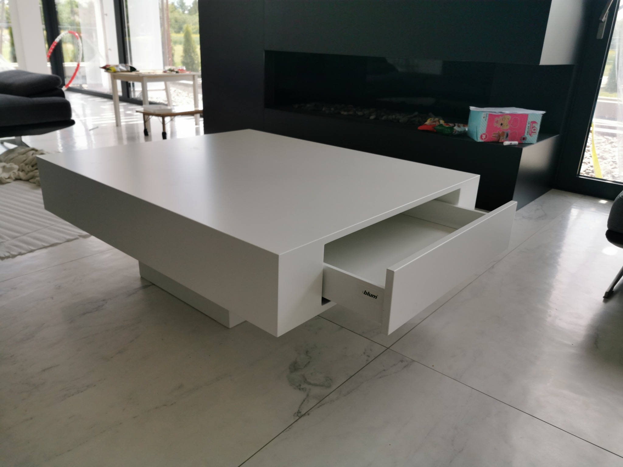 Nowoczesny, biały stolik kawowy z wysuwaną szufladą na tle jasnej podłogi z marmurkowym wzorem i minimalistycznego kominka.