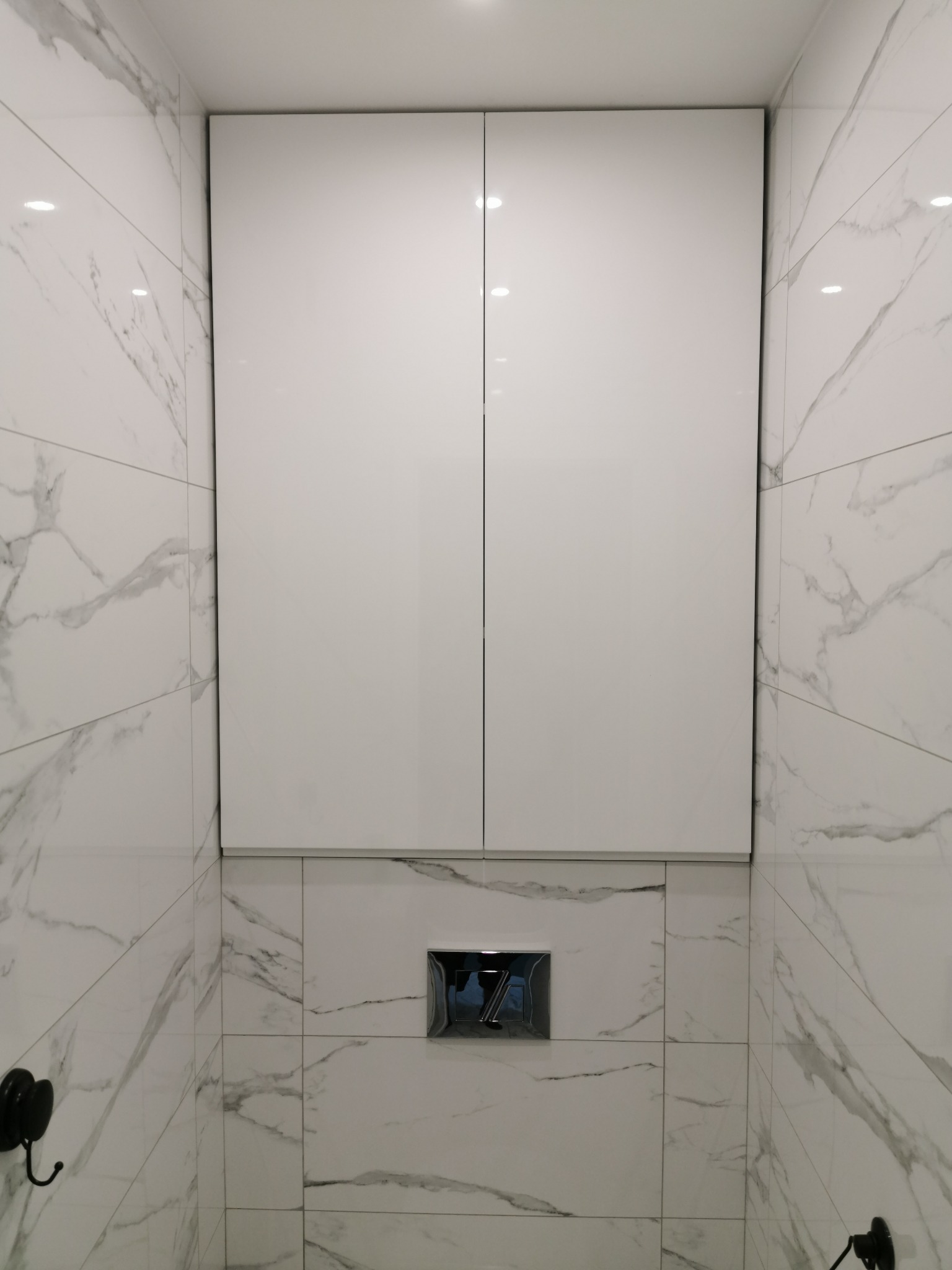 Biała szafka wisząca nad toaletą, zintegrowana ze ścianą wyłożoną marmurowymi płytkami, chromowany przycisk spłuczki w tle.