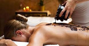 Kobieta leżąca na brzuchu podczas zabiegu czekoladowego na plecy. Terapeuta aplikuje ciepłą, płynną czekoladę z czarnego pojemnika. W tle świecą się rozproszone światła świec.