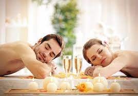 Para relaksuje się na stole, przygotowanym do zabiegu, z zapalonymi świecami, kwiatami i szampanem, oboje mają zamknięte oczy, kobieta ma kwiat we włosach.