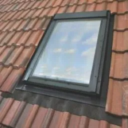 Okno dachowe wkomponowane w czerwoną dachówkę, widoczne niebo z lekkimi chmurami odbijające się w szybie.