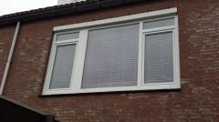 Białe okno PCV z żaluzjami poziomymi zamontowane w ścianie z czerwonej cegły. Widoczne fragmenty dachu i budynku w tle.