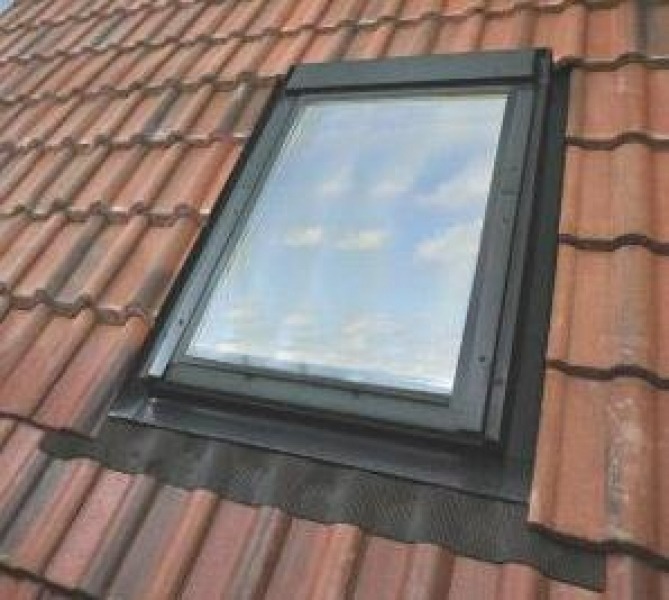 Okno dachowe wkomponowane w czerwoną dachówkę, widoczne niebo z lekkimi chmurami odbijające się w szybie.
