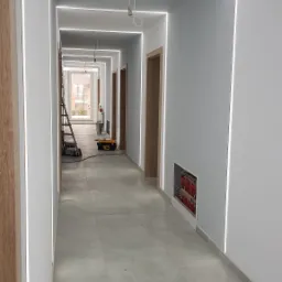 Długi korytarz w trakcie prac wykończeniowych, z zamontowanym oświetleniem LED wzdłuż ścian i sufitu, widoczne narzędzia i drabina w tle, surowe drewniane futryny drzwiowe, szara podłoga z płytek.