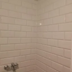 Białe, błyszczące, prostokątne płytki ułożone w cegiełkę na ścianie w rogu łazienki, z widoczną chromowaną baterią umywalkową.