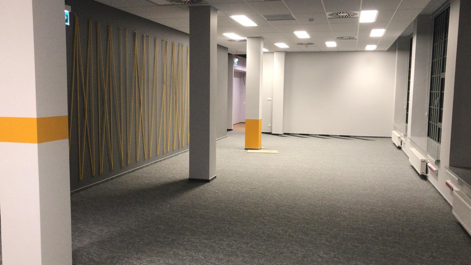 Nowoczesne biuro po remoncie: szara wykładzina, białe ściany, żółte akcenty na kolumnach i dekoracyjne sznury na ścianie, widoczne grzejniki pod oknami.