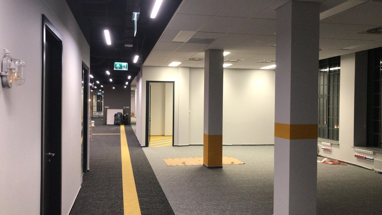 Wnętrze biurowe w trakcie wykańczania: szary dywan w pasy, pomalowane filary z żółtymi akcentami, lampy sufitowe, drzwi pomalowane na czarno, lampa w stylu industrialnym na ścianie, widoczne kable...