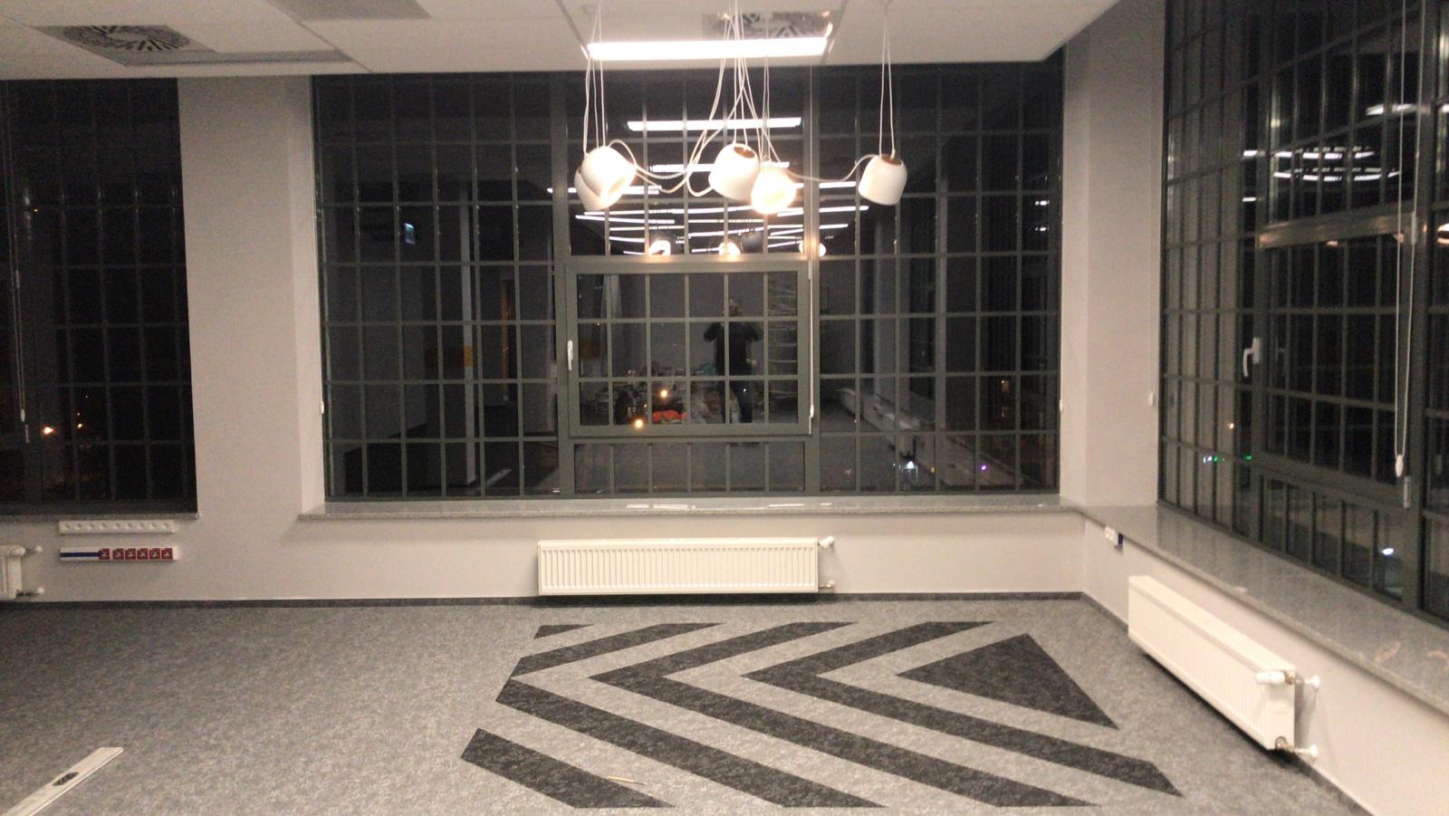 Pusta przestrzeń biurowa z dużymi oknami w stylu industrialnym, wzorzysta wykładzina podłogowa w zygzaki i designerska lampa wisząca z ceramicznymi kloszami.