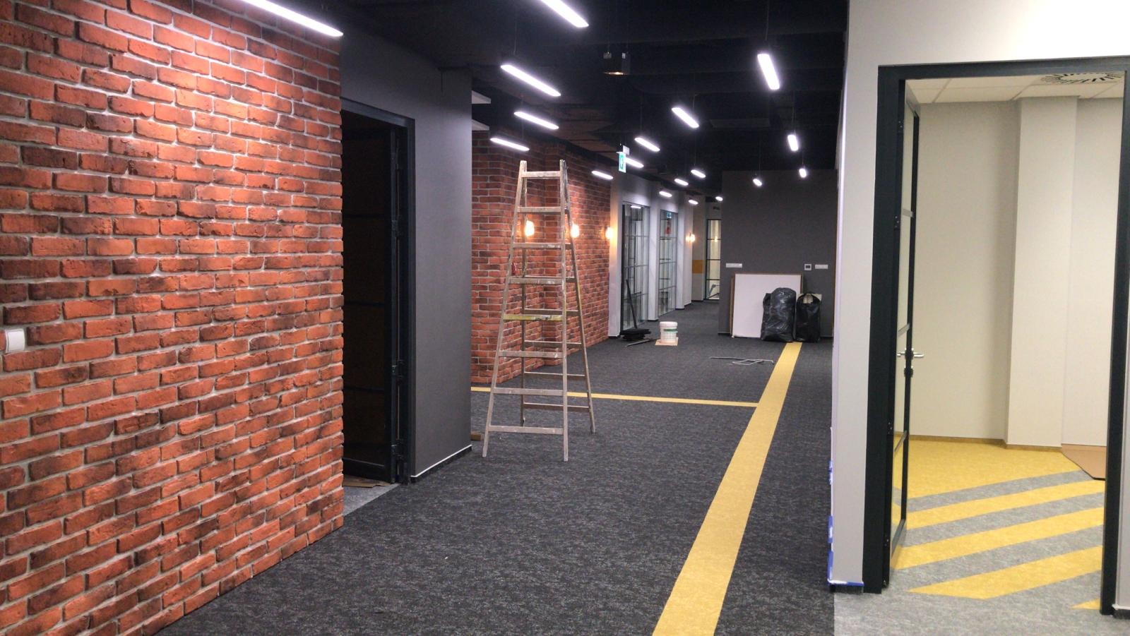 Korytarz biurowy w trakcie wykańczania: ściana z cegły, drabina, czarna podłoga z żółtymi pasami, widoczne prace wykończeniowe.
