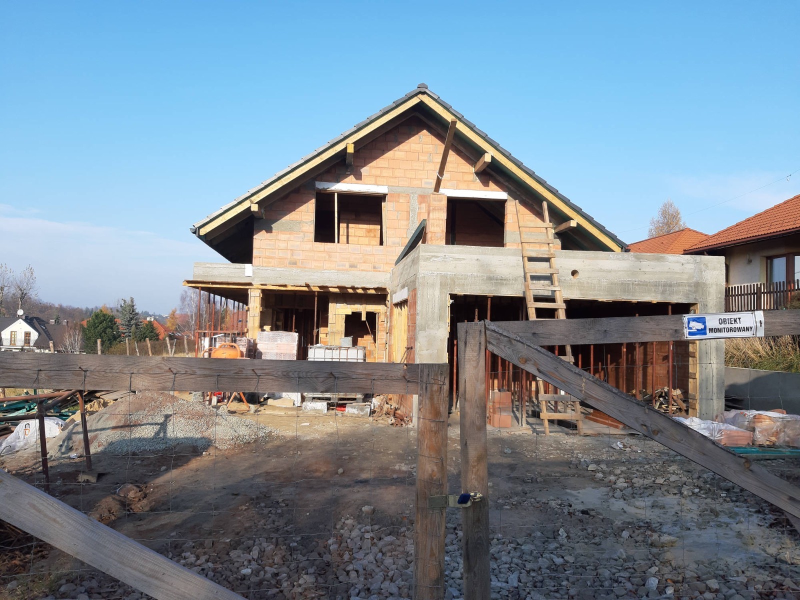 Budowa domu jednorodzinnego w stanie surowym otwartym, widoczne ściany z czerwonej cegły, betonowe elementy konstrukcyjne, drewniana drabina oparta o ścianę, teren budowy ogrodzony drewnianym...