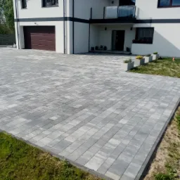 Ułożona szara kostka brukowa na podjeździe przed nowoczesnym domem z brązową bramą garażową i szklaną balustradą balkonu, widoczne donice z roślinami.