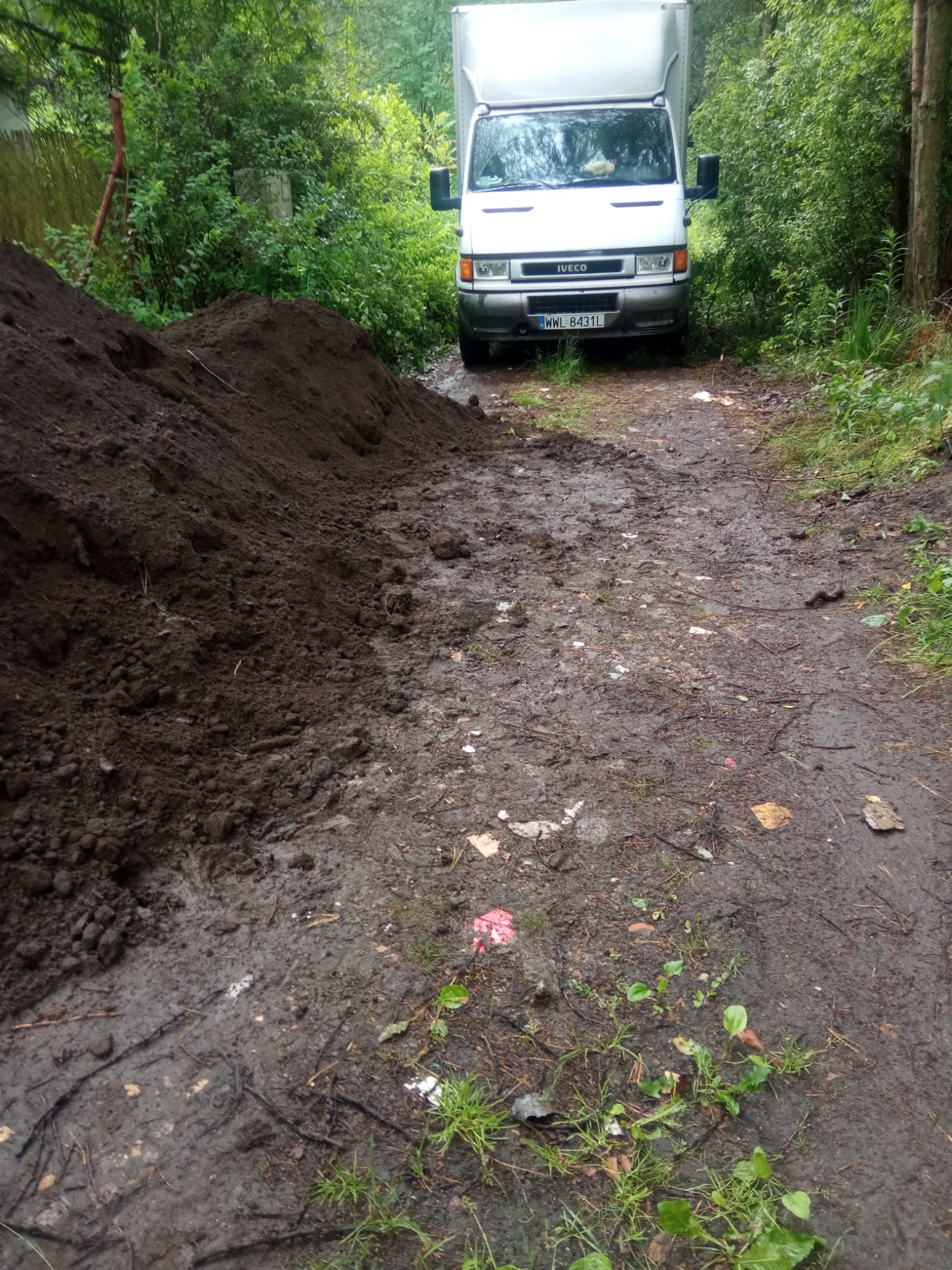 Stos brudnej ziemi obok błotnistej drogi dojazdowej z białym samochodem dostawczym Iveco w tle.