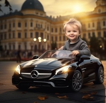 Uśmiechnięty blond chłopiec siedzi za kierownicą czarnego, elektrycznego samochodu-zabawki, imitującego Mercedesa, na tle rozmytego, klasycystycznego budynku oświetlonego zachodzącym słońcem.