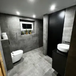 Nowoczesna łazienka z szarymi płytkami, wiszącą toaletą, czarną szafką z białą umywalką i oknem z roślinami w doniczkach.