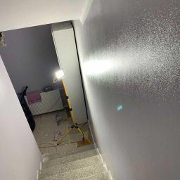 BenBud - Klatka schodowa w trakcie prac wykończeniowych, świeżo pomalowana ściana z efektem dekoracyjnym, widoczny reflektor budowlany oświetlający powierzchnię, schody zabezpieczone folią malarską.