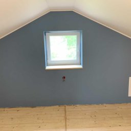 BenBud - Pomalowane na szaro poddasze ze skośnym sufitem, widoczne okno dachowe z drewnianym parapetem i fragment drewnianej podłogi przed położeniem listew przypodłogowych.