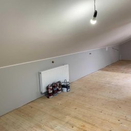 BenBud - Pomalowane na szaro poddasze ze skośnym sufitem, drewnianą podłogą i grzejnikiem przy ścianie. Kilka puszek z farbą stoi obok grzejnika.