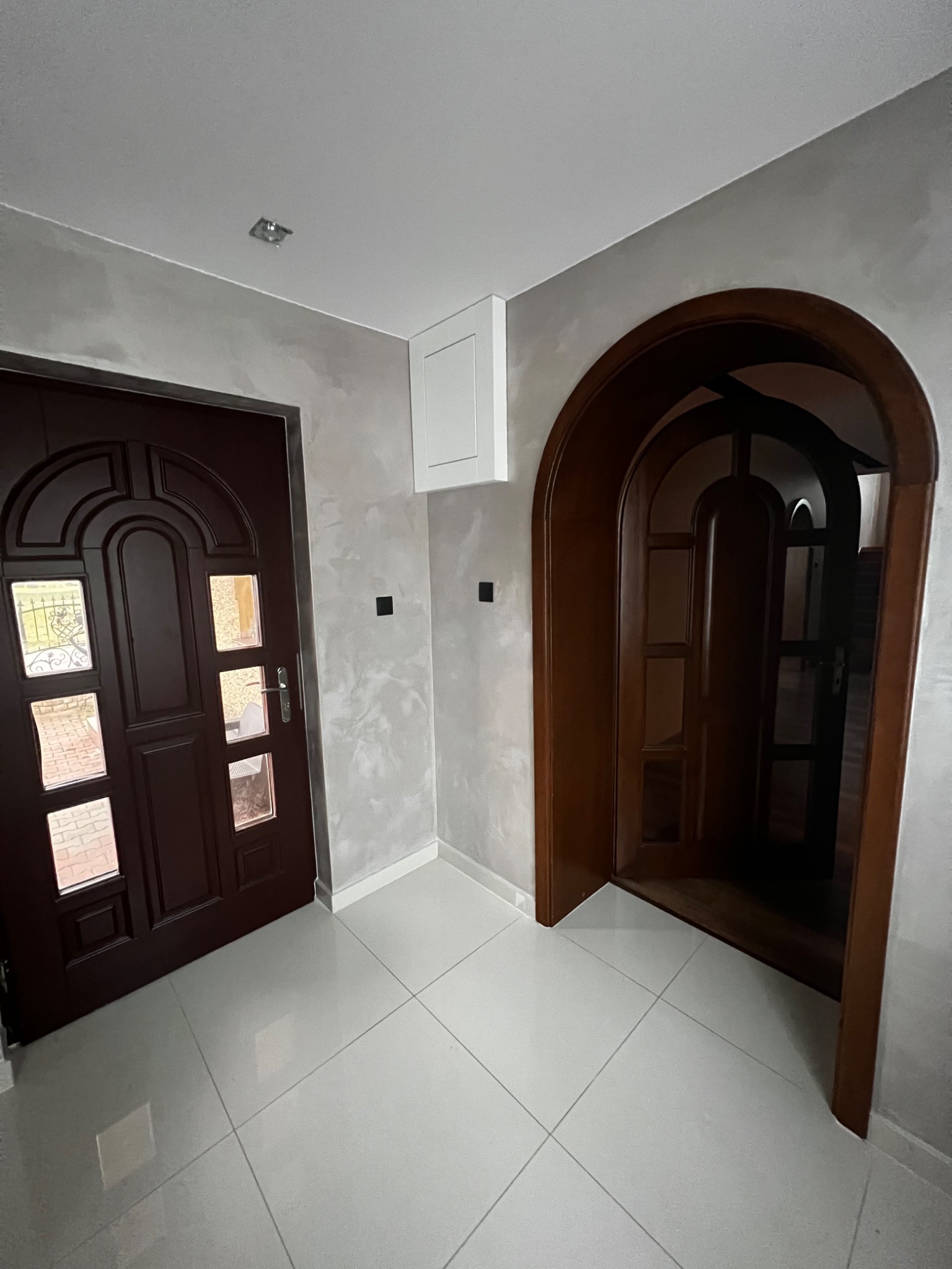 Wnętrze domu z dwoma ciemnobrązowymi drzwiami, jedne standardowe z szybkami, drugie w formie łukowatego przejścia, na podłodze białe płytki, ściany w odcieniach szarości z efektem dekoracyjnym.