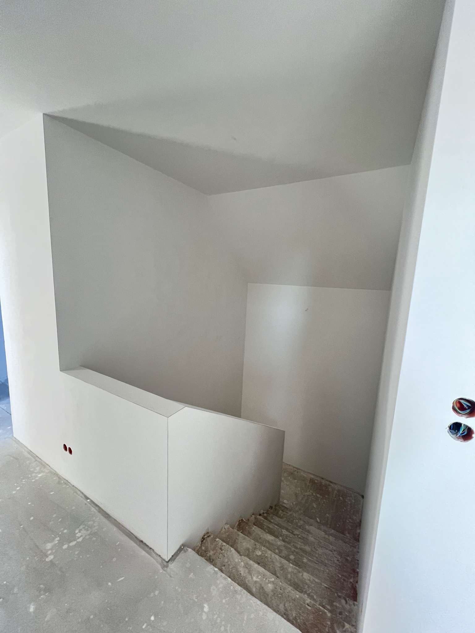 Klatka schodowa w trakcie wykańczania, ściany i sufit pomalowane na biało, widoczne niedoskonałości na schodach i podłodze, instalacja elektryczna wystaje ze ściany.