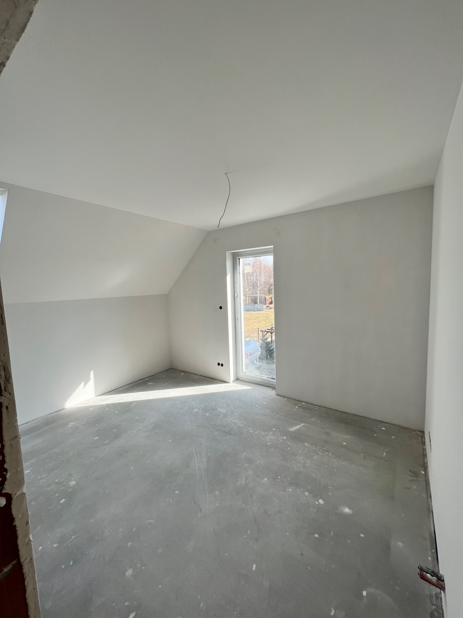 Pusty pokój po świeżo wykonanych pracach wykończeniowych, ściany pomalowane na biało, surowa betonowa podłoga, widoczne ślady po pracach, okno z widokiem na zewnątrz.