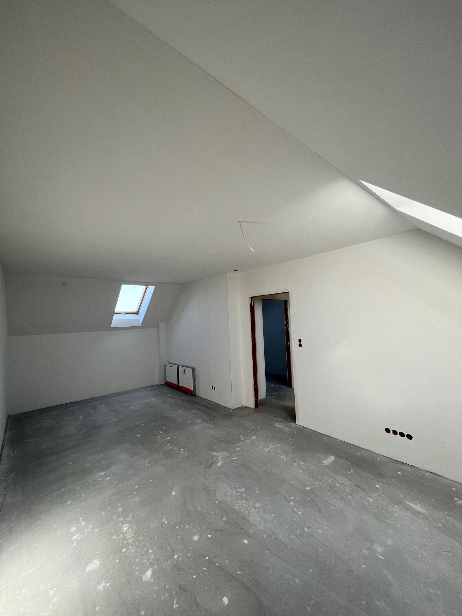 Poddasze po remoncie: białe ściany, surowe wylewki na podłodze, okno dachowe, widoczne puszki elektryczne i futryny drzwiowe.