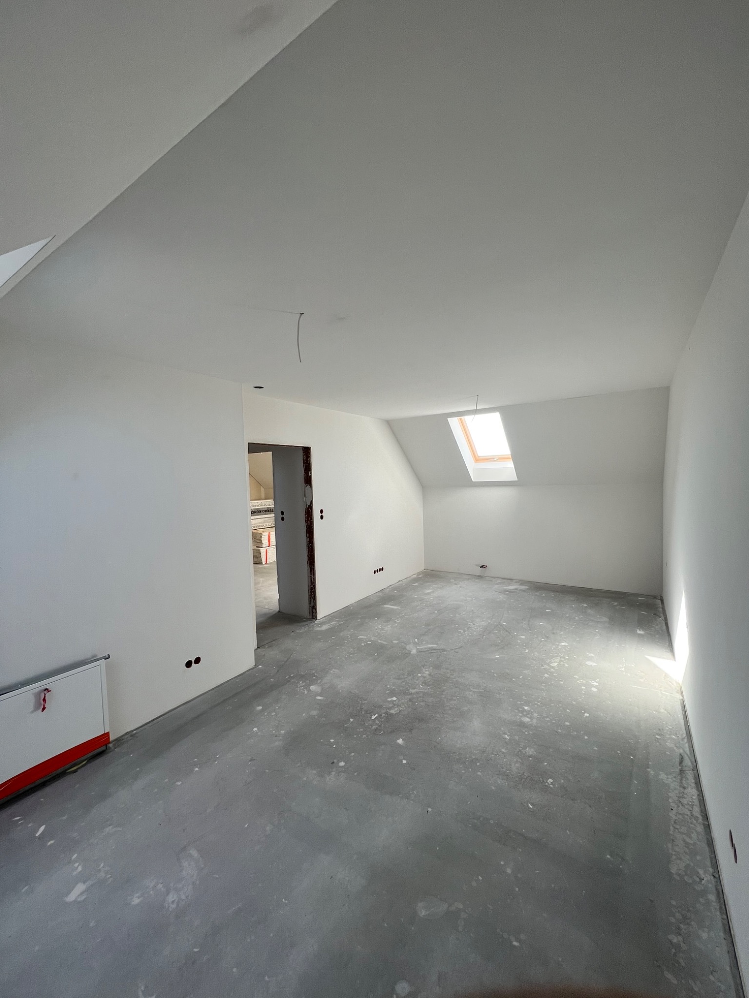 Poddasze po świeżym malowaniu na biało, widoczne ślady po pracach na szarym betonie, okno dachowe wpuszczające jasne światło, przygotowane do dalszych prac wykończeniowych.