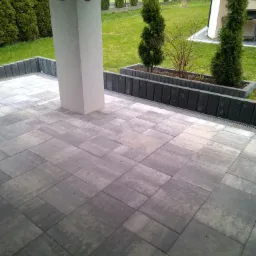 Nowoczesny taras z szarych płyt betonowych, obrzeża z kostki, donice z tujami i krzewami, widok na trawnik i dom w tle.