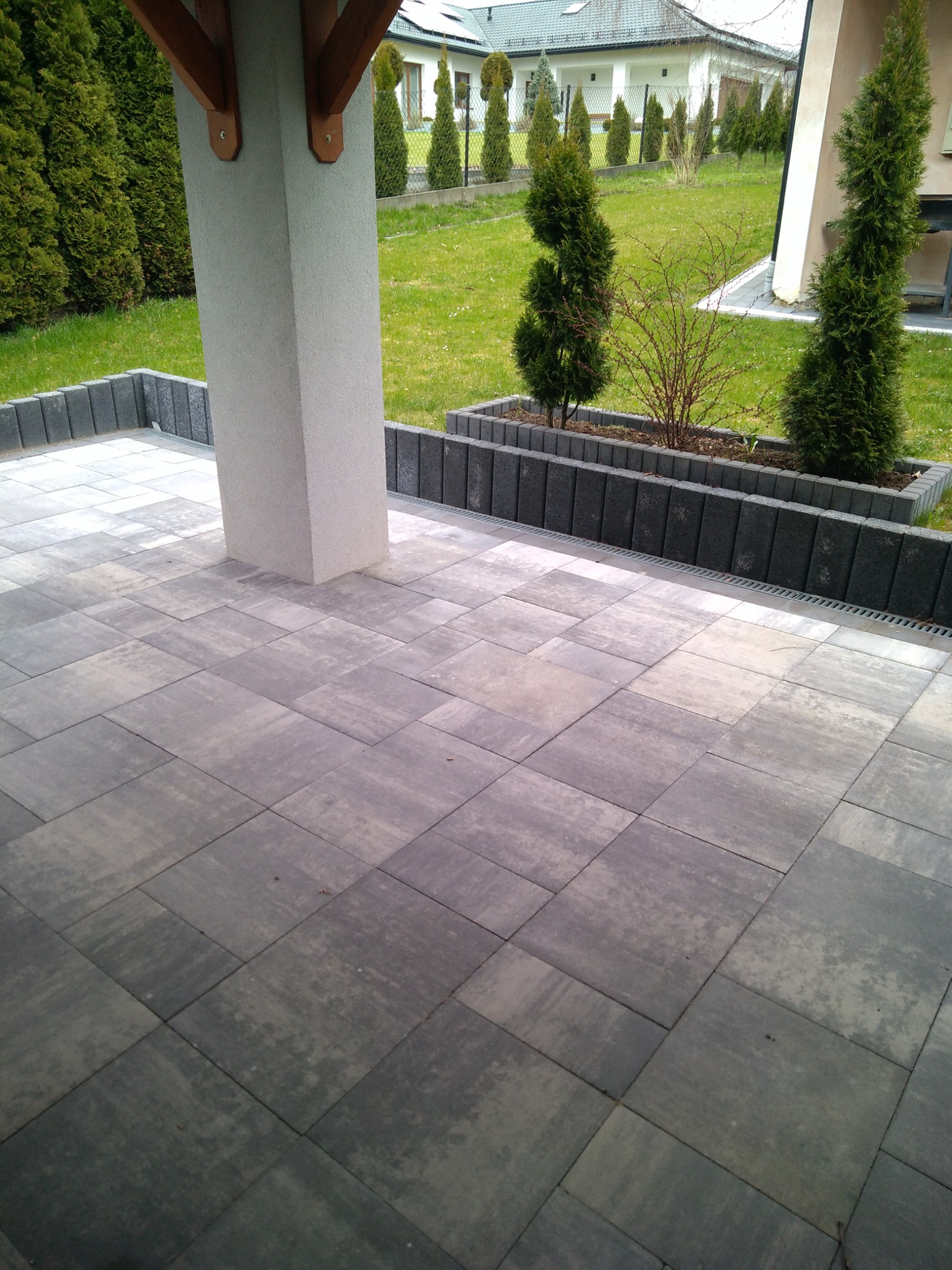 Nowoczesny taras z szarych płyt betonowych, obrzeża z kostki, donice z tujami i krzewami, widok na trawnik i dom w tle.