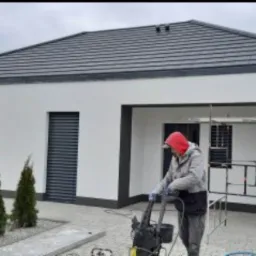 Pracownik w czerwonej bluzie myje elewację nowoczesnego, parterowego domu z szarą stolarką okienną i ciemną dachówką przy użyciu myjki ciśnieniowej. Na drugim planie widoczne rusztowanie i wiaderka.