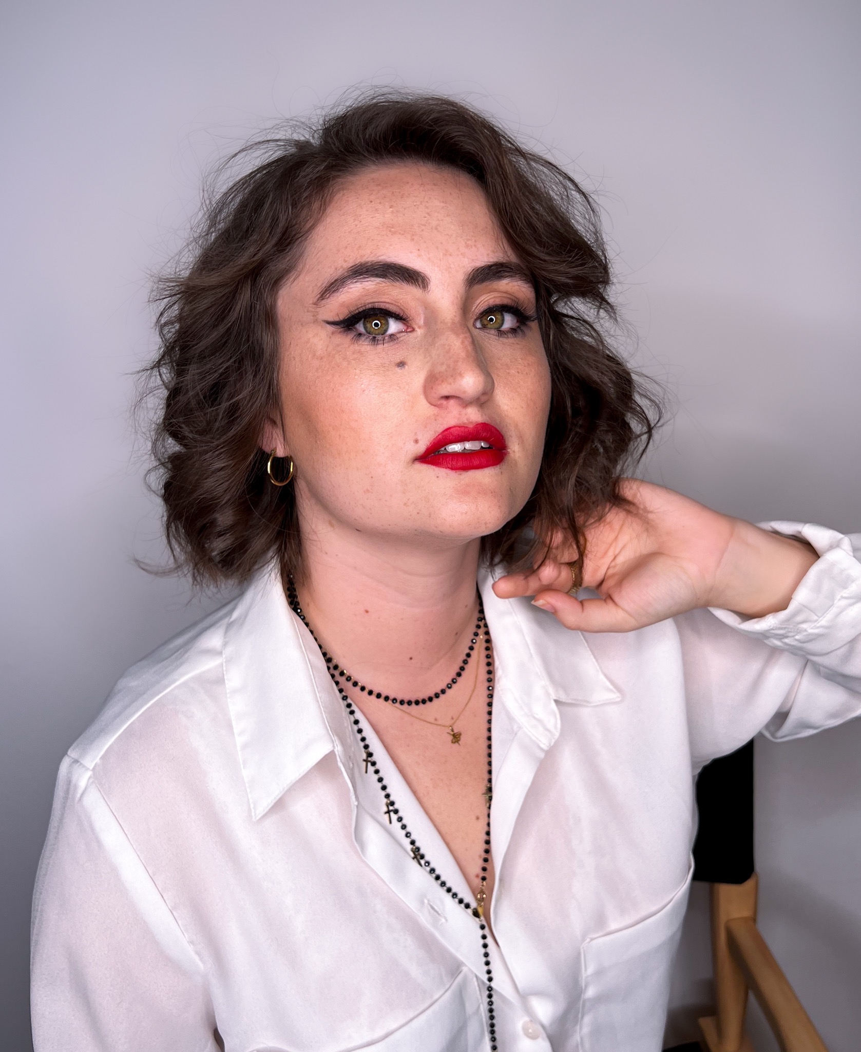 Portret kobiety z krótkimi, kręconymi włosami, wyrazistą czerwoną szminką i czarnym eyelinerem, pozującej w białej koszuli i z delikatną biżuterią na szyi.