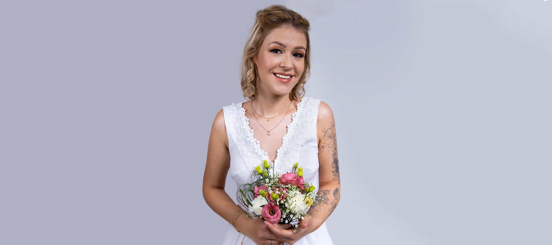Uśmiechnięta blondynka z upiętymi włosami, delikatnym makijażem i tatuażami na ramieniu, trzymająca bukiet różowych, białych i zielonych kwiatów na jasnoszarym tle.