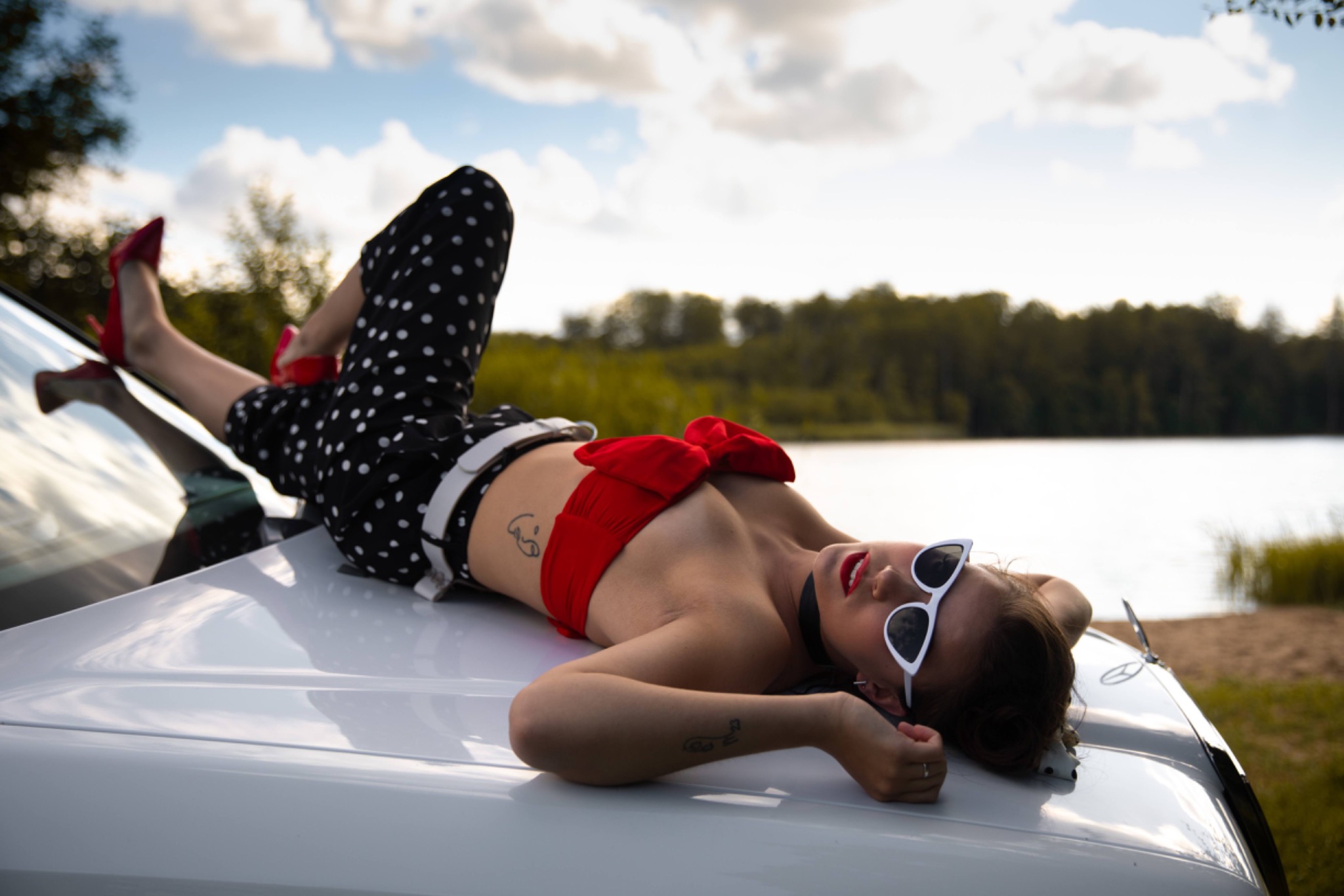 Kobieta w stylu retro leży na białym samochodzie, w czerwonym topie i okularach, z czerwonymi ustami, na tle jeziora i zieleni.
