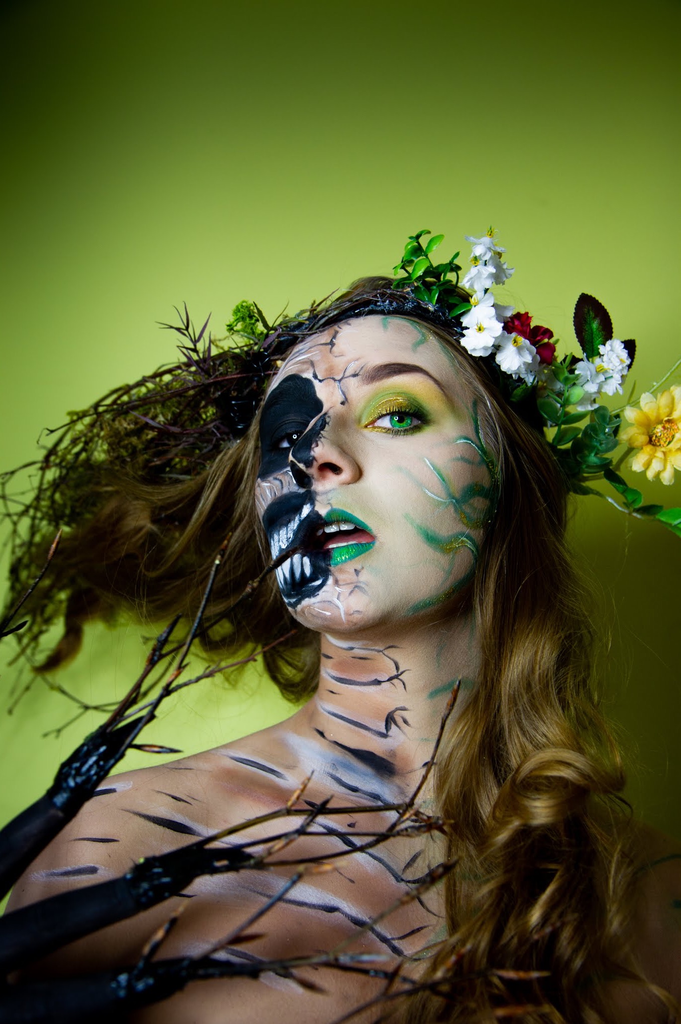 Kobieta z artystycznym body paintingiem i makijażem, połowa twarzy w motywie czaszki, druga połowa z zielonymi akcentami i kwiatowym wiankiem na głowie, na jednolitym zielonym tle.