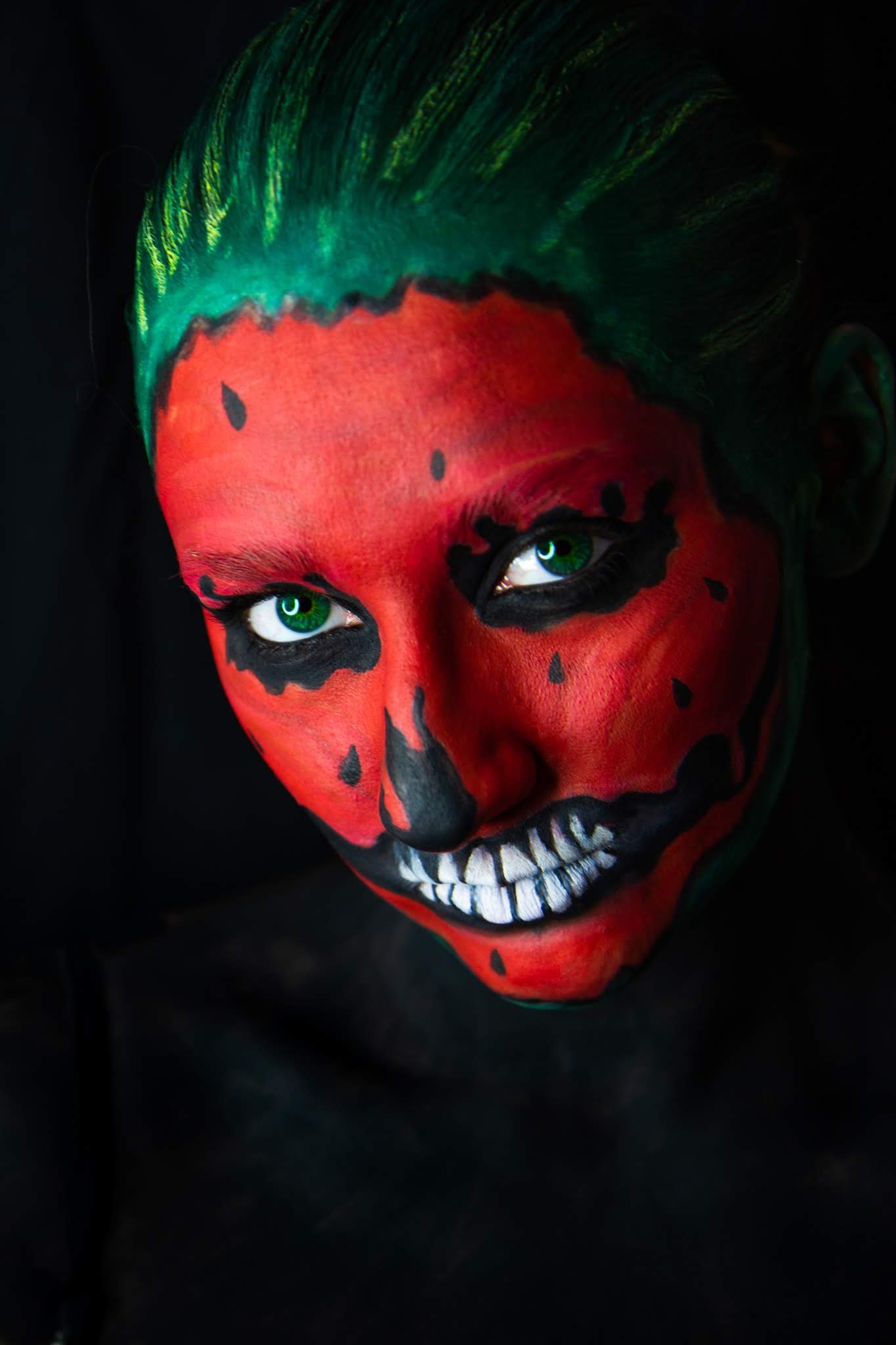 Portret kobiety z awangardowym makijażem: czerwona twarz z czarnymi plamkami imitującymi arbuza, zielone włosy i intensywne, zielone oczy spoglądające w dół, na ciemnym tle.