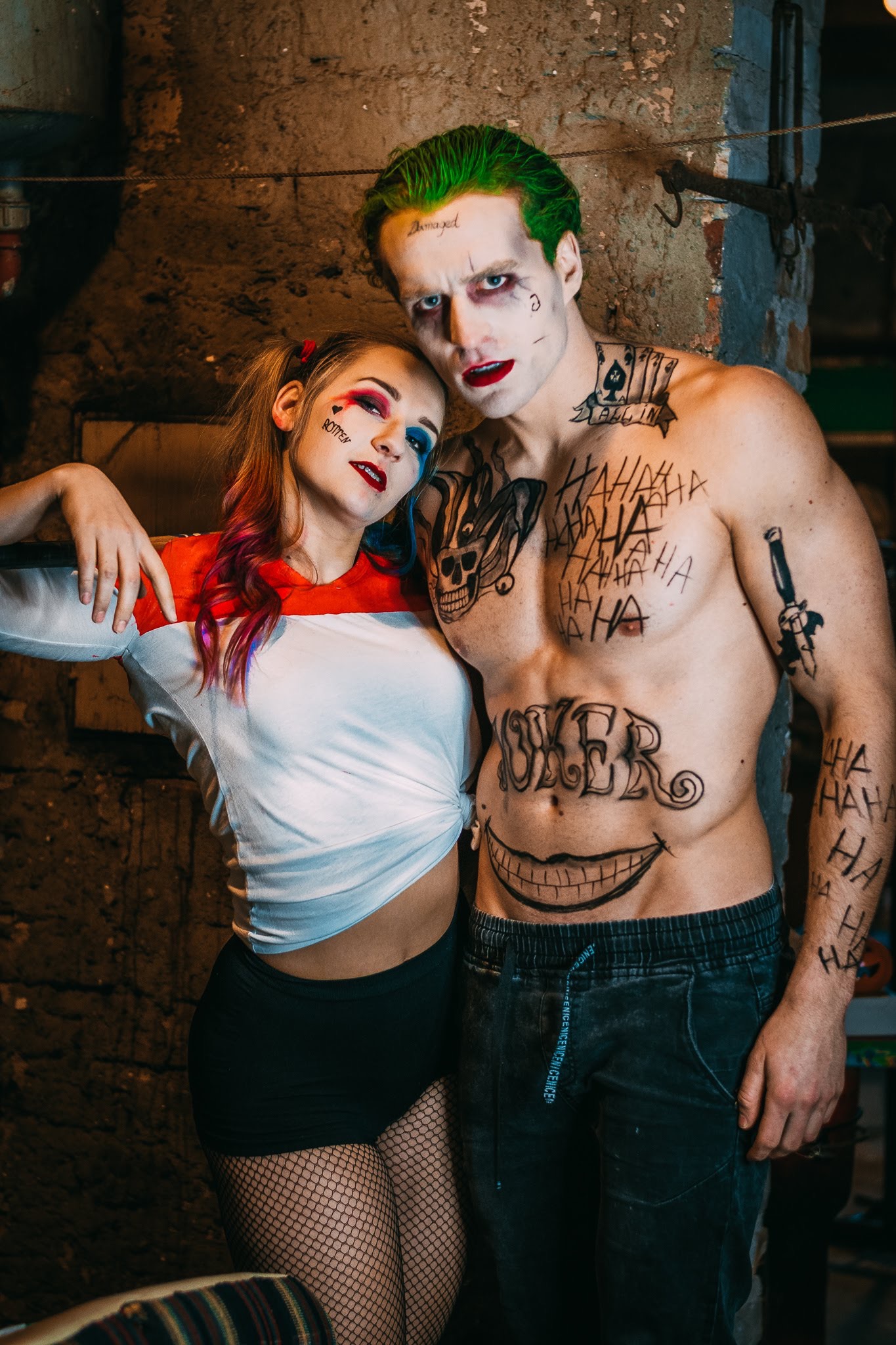 Para w stylizacjach Harley Quinn i Jokera: kobieta z czerwono-niebieskim makijażem oczu i serduszkiem na policzku, mężczyzna z zielonymi włosami, białym makijażem, czerwonymi ustami i napisem...