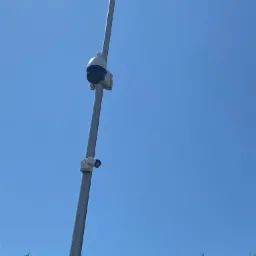 Dwie kamery monitoringu, jedna obrotowa PTZ, zamontowane na wysokim, szarym słupie na tle błękitnego nieba i wierzchołków zielonych drzew.