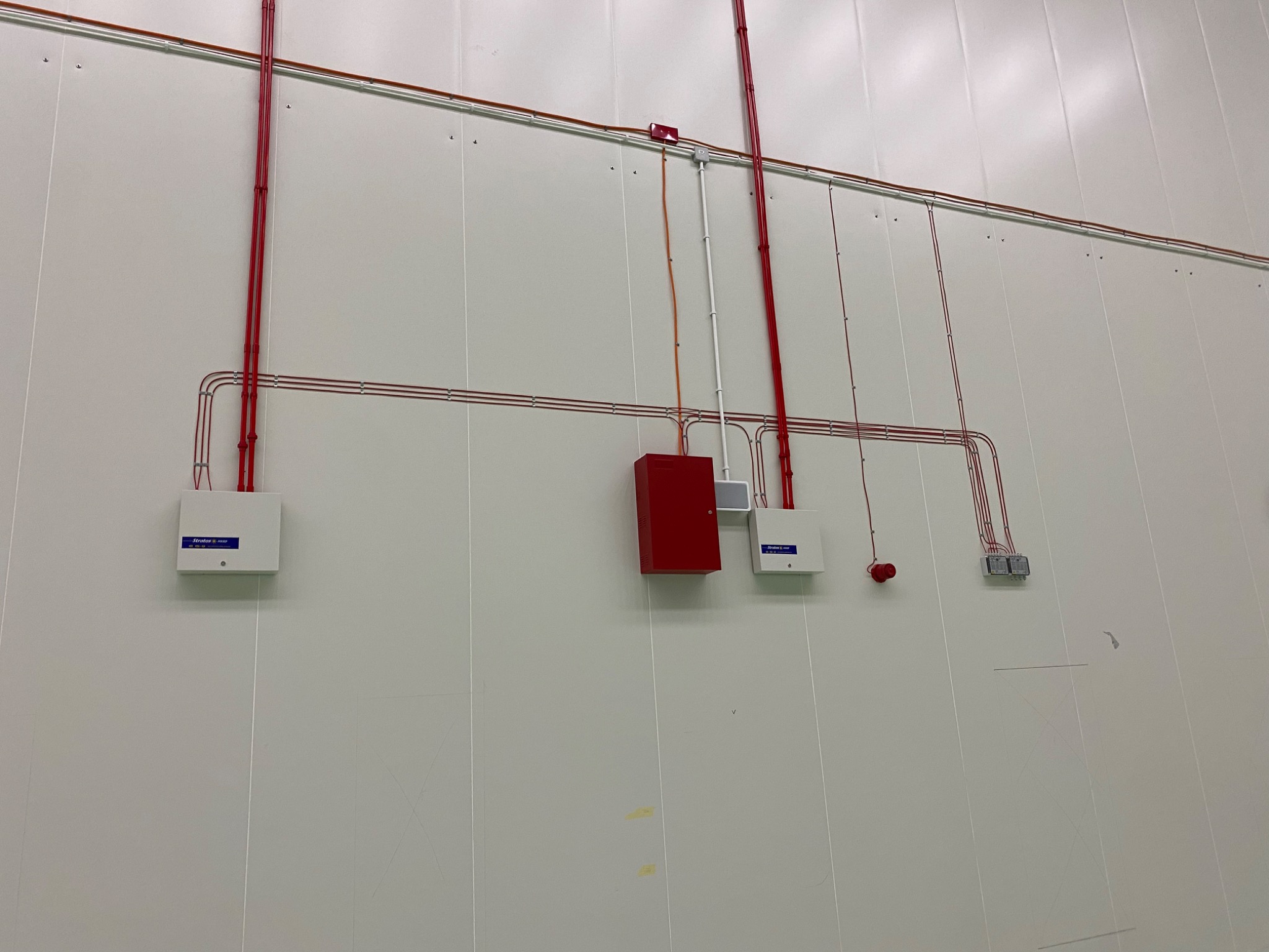 Biała ściana z panelami, na której zamontowano instalacje elektryczne w postaci przewodów w czerwonej osłonie, białych i czerwonych puszek oraz czujnika dymu
