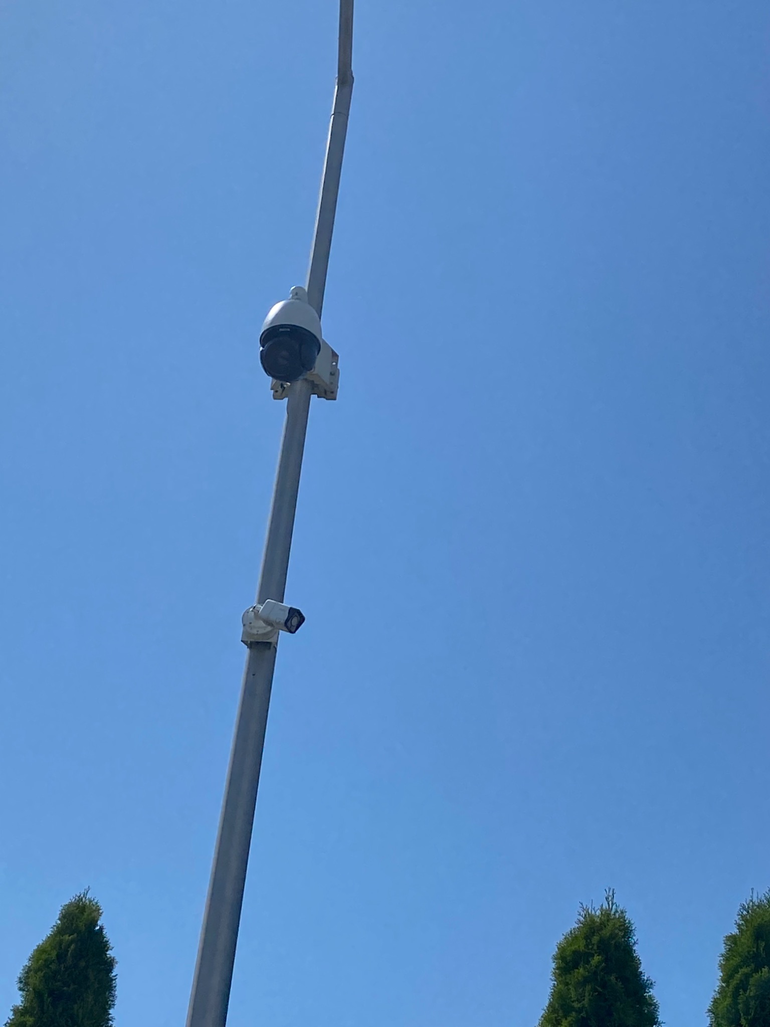 Dwie kamery monitoringu, jedna obrotowa PTZ, zamontowane na wysokim, szarym słupie na tle błękitnego nieba i wierzchołków zielonych drzew.