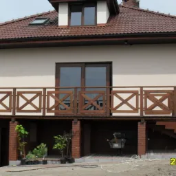 Dwukondygnacyjny dom z beżową elewacją, brązowym dachem i balkonem z drewnianą balustradą w kształcie krzyżyków, widok z zewnątrz.