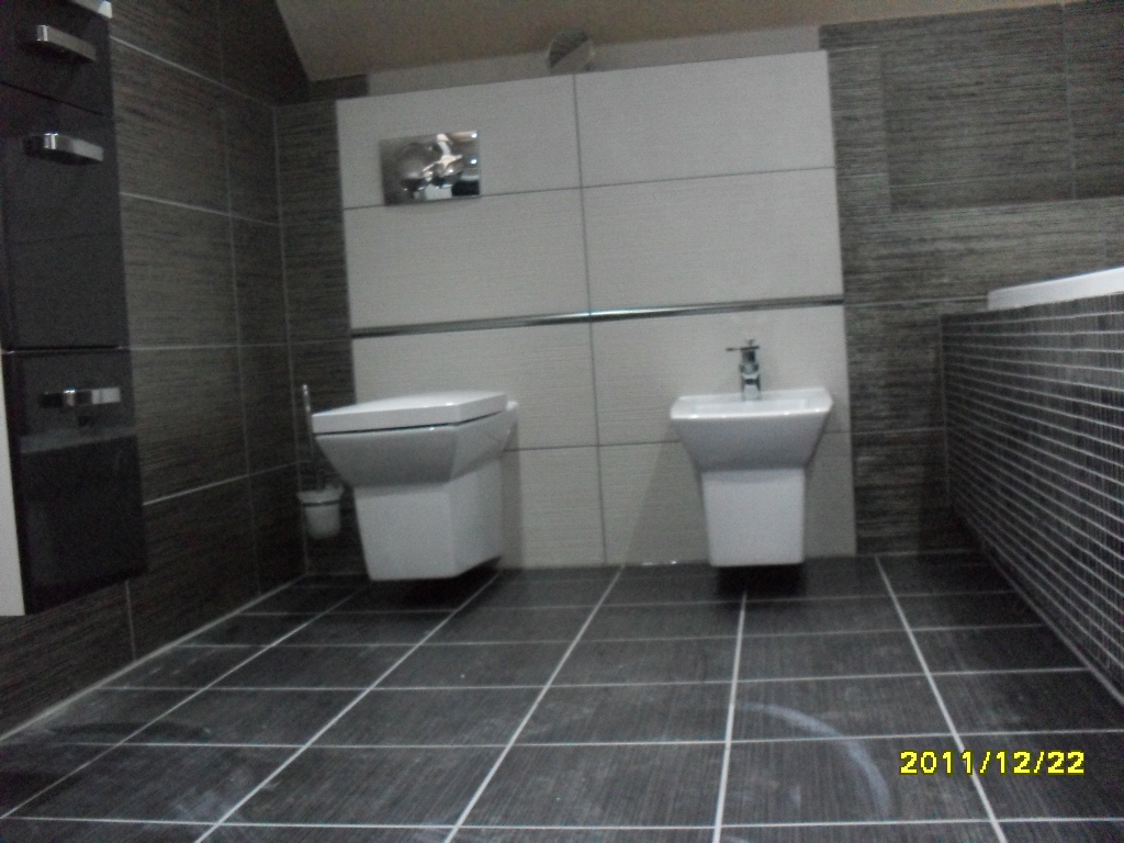 Wykończona łazienka z ciemnymi płytkami podłogowymi i ściennymi, wisząca toaleta i umywalka na postumencie, białe płytki na ścianie za urządzeniami, data na zdjęciu w prawym dolnym rogu.