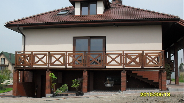 Dwukondygnacyjny dom z beżową elewacją, brązowym dachem i balkonem z drewnianą balustradą w kształcie krzyżyków, widok z zewnątrz.