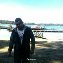 Mężczyzna w kurtce na tle jeziora z pomostem i budynkiem w oddali.