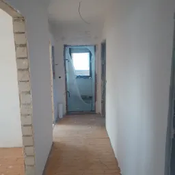 Korytarz w trakcie prac wykończeniowych: ściany po nałożeniu gładzi, widoczne fragmenty surowej cegły przy ościeżnicach, drewniana podłoga częściowo zakryta folią ochronną, okno zasłonięte folią...