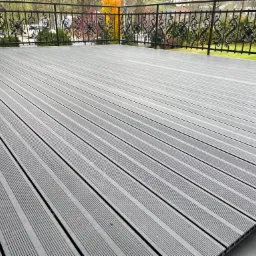 Szara deska kompozytowa tarasu ułożona równolegle, widok z góry, czarna, ozdobna balustrada z metalu w tle, widoczne jesienne drzewa.