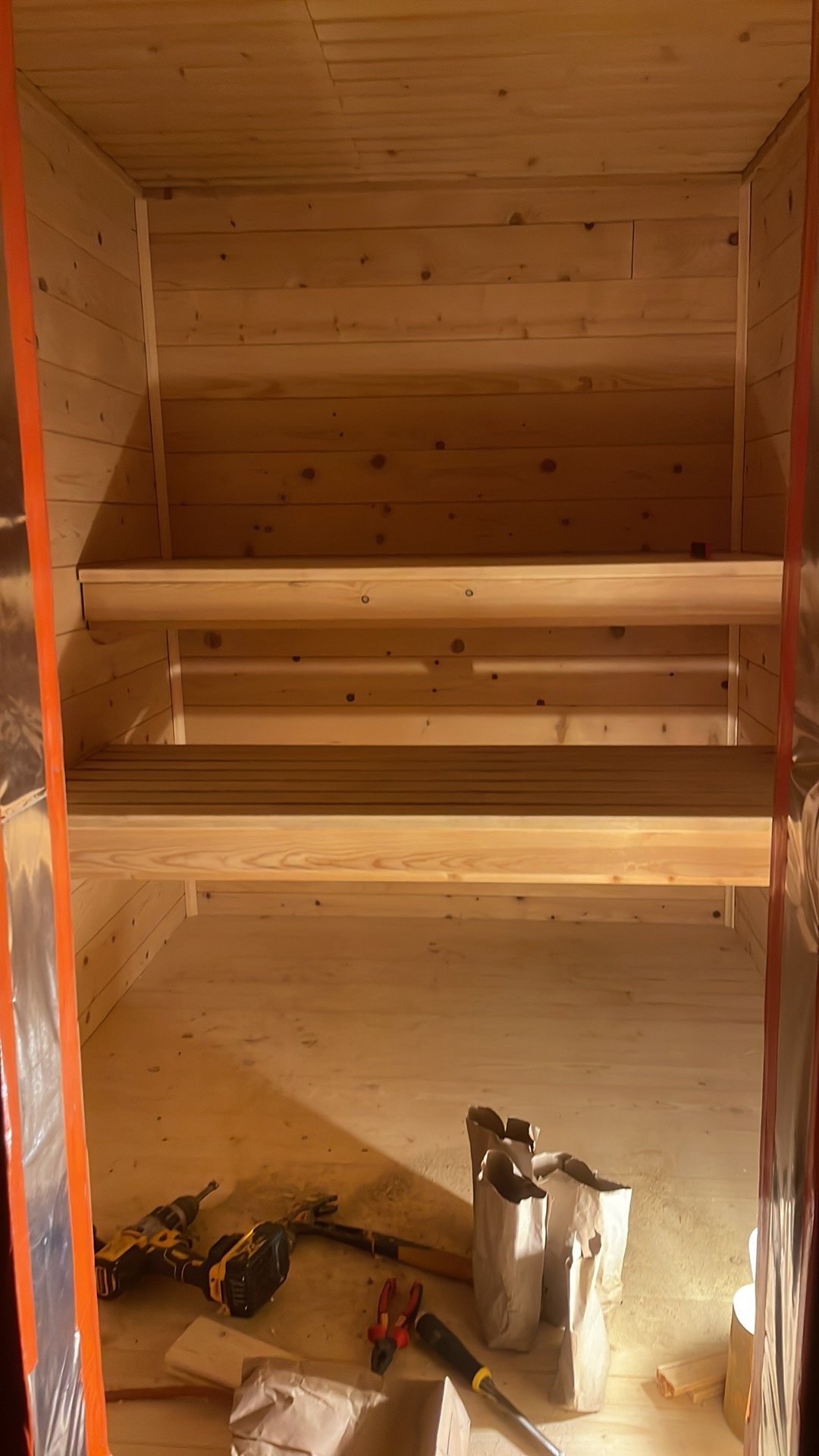 Wnętrze sauny w trakcie budowy z widocznymi drewnianymi ławkami, narzędziami (wiertarka, młotek, kombinerki, śrubokręt) i workami z materiałami na podłodze, zabezpieczone folią ściany.