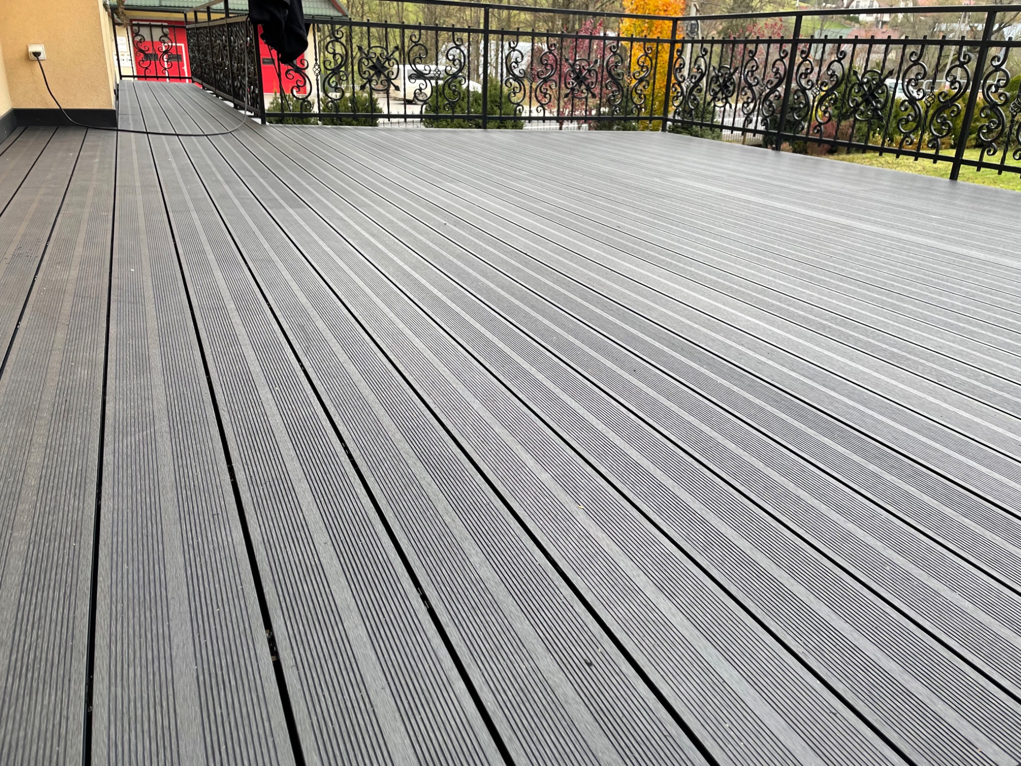 Szara deska kompozytowa na tarasie z ozdobną, kutą balustradą, widok na ogród z drzewami o jesiennych barwach.