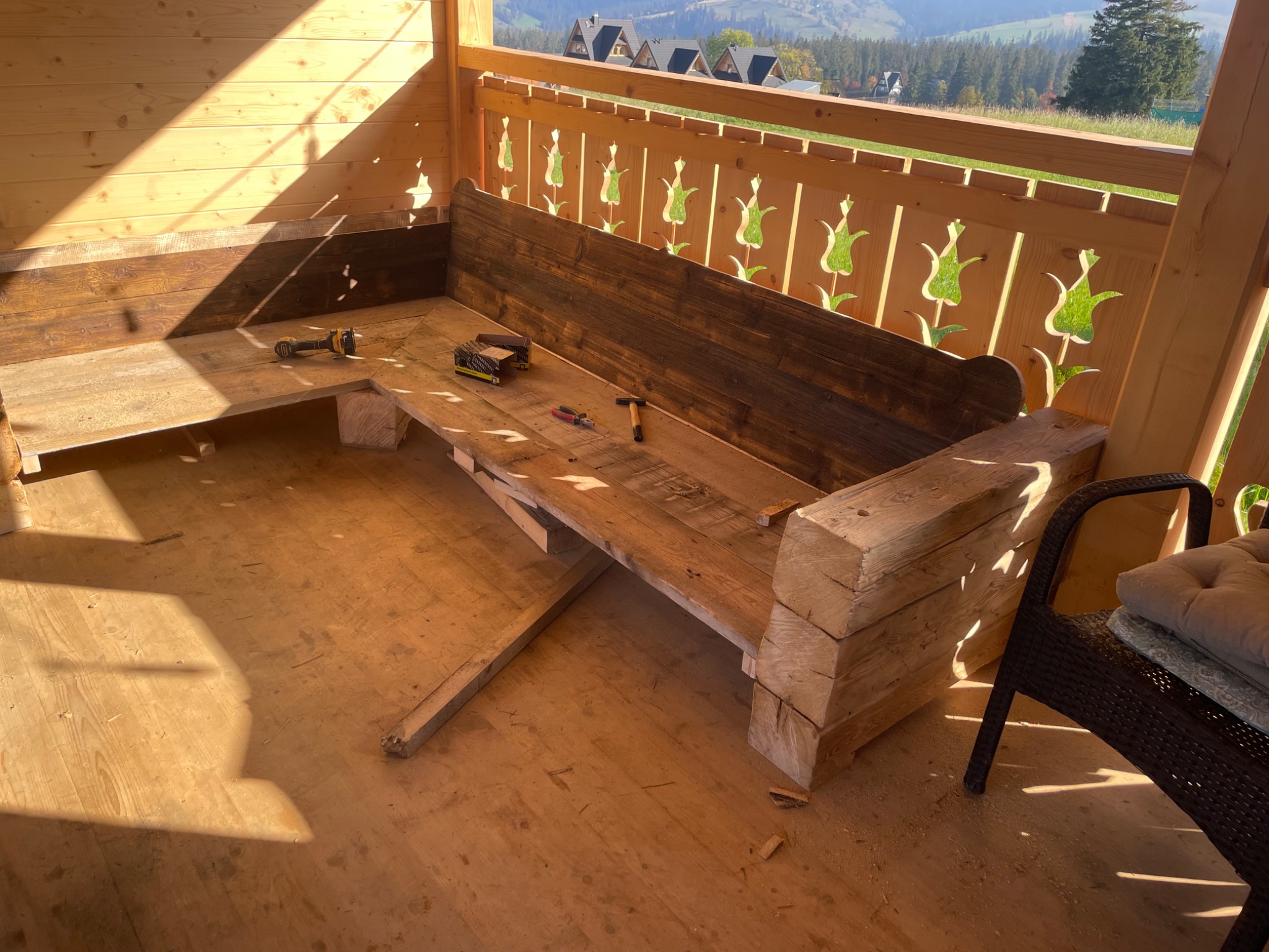 Wnętrze drewnianej altany z częściowo wykonaną ławką z grubych bali, narzędzia leżą na siedzisku, w tle widok na górskie zbocza.