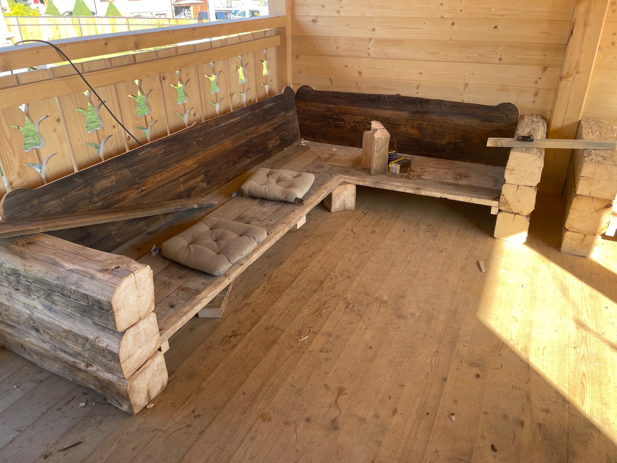 Narożna ławka z surowego drewna z poduszkami w altanie, widoczne narzędzia i elementy konstrukcyjne w trakcie montażu.