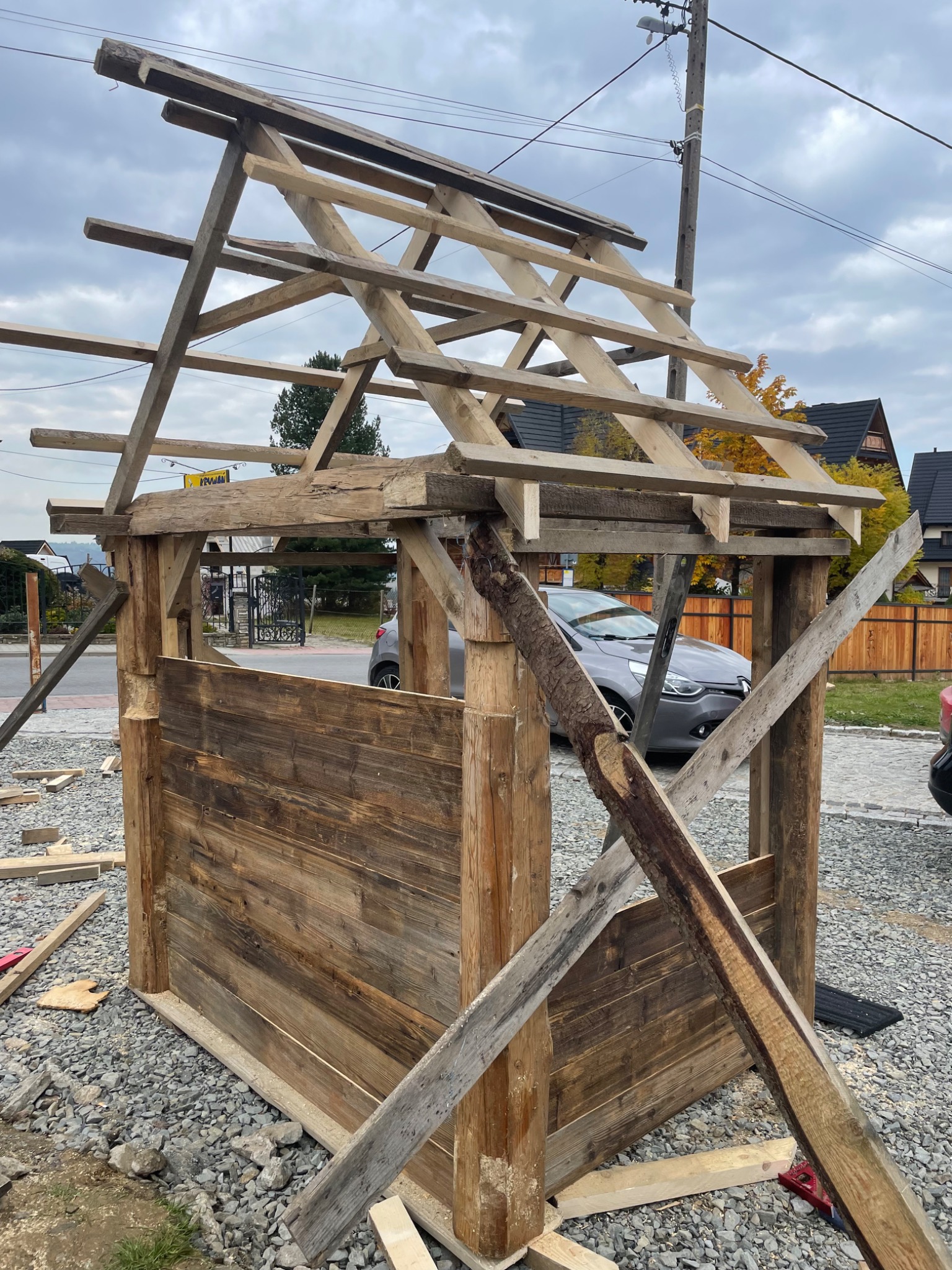 Konstrukcja altany z surowego drewna w trakcie budowy, widoczne deski ścian i drewniany szkielet dachu na tle szarej nawierzchni i zabudowań.